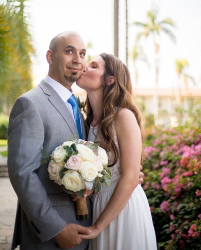 Bride kissing the groom's cheek in front of pink bougainvillea and palms