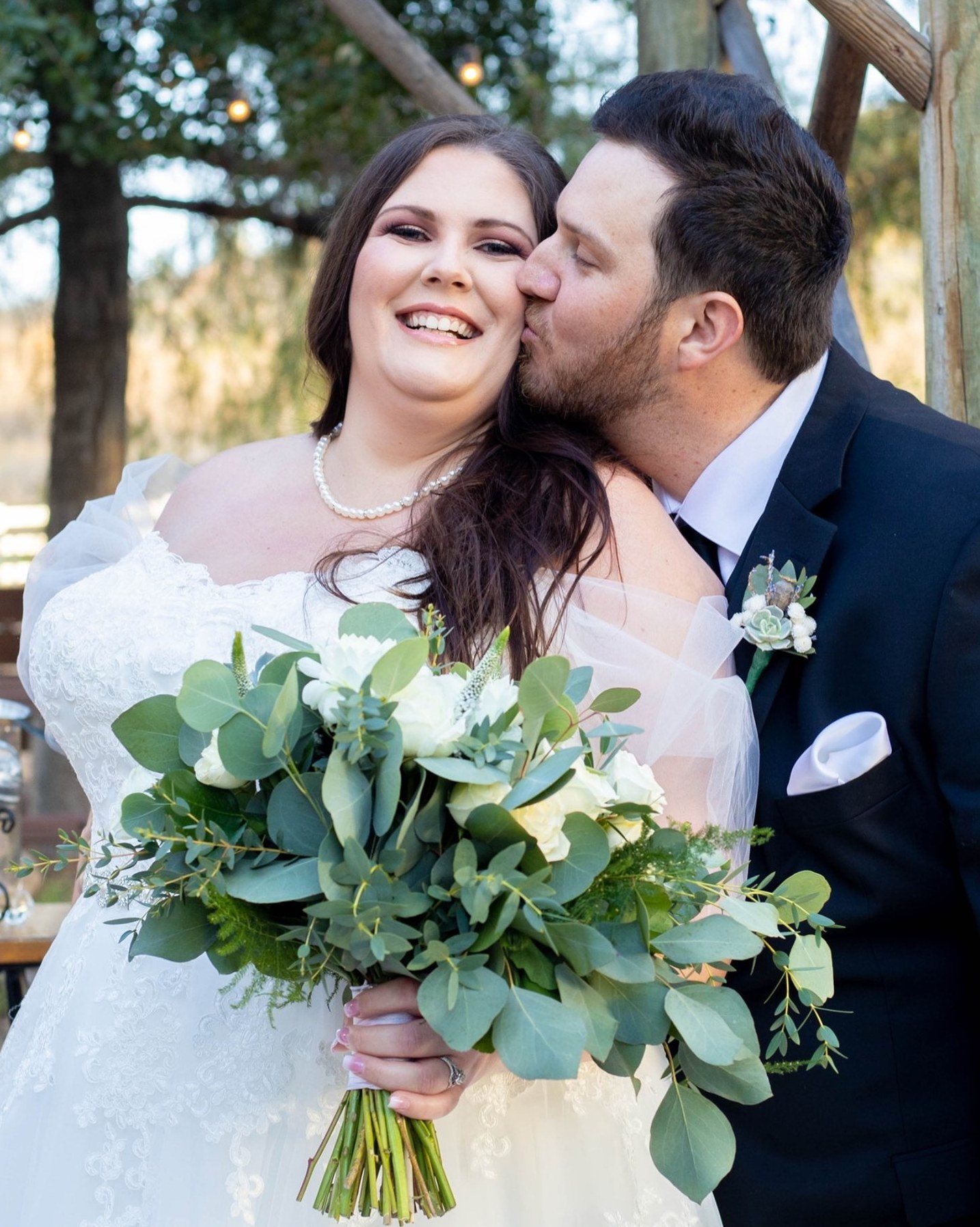 Bride laughing while the groom kisses her cheek, eucalyptus bouquet in hand