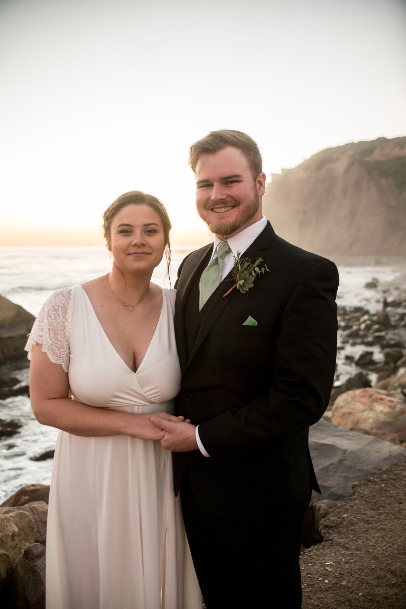 Bride and groom smiling on the coast below a sunlit bluff