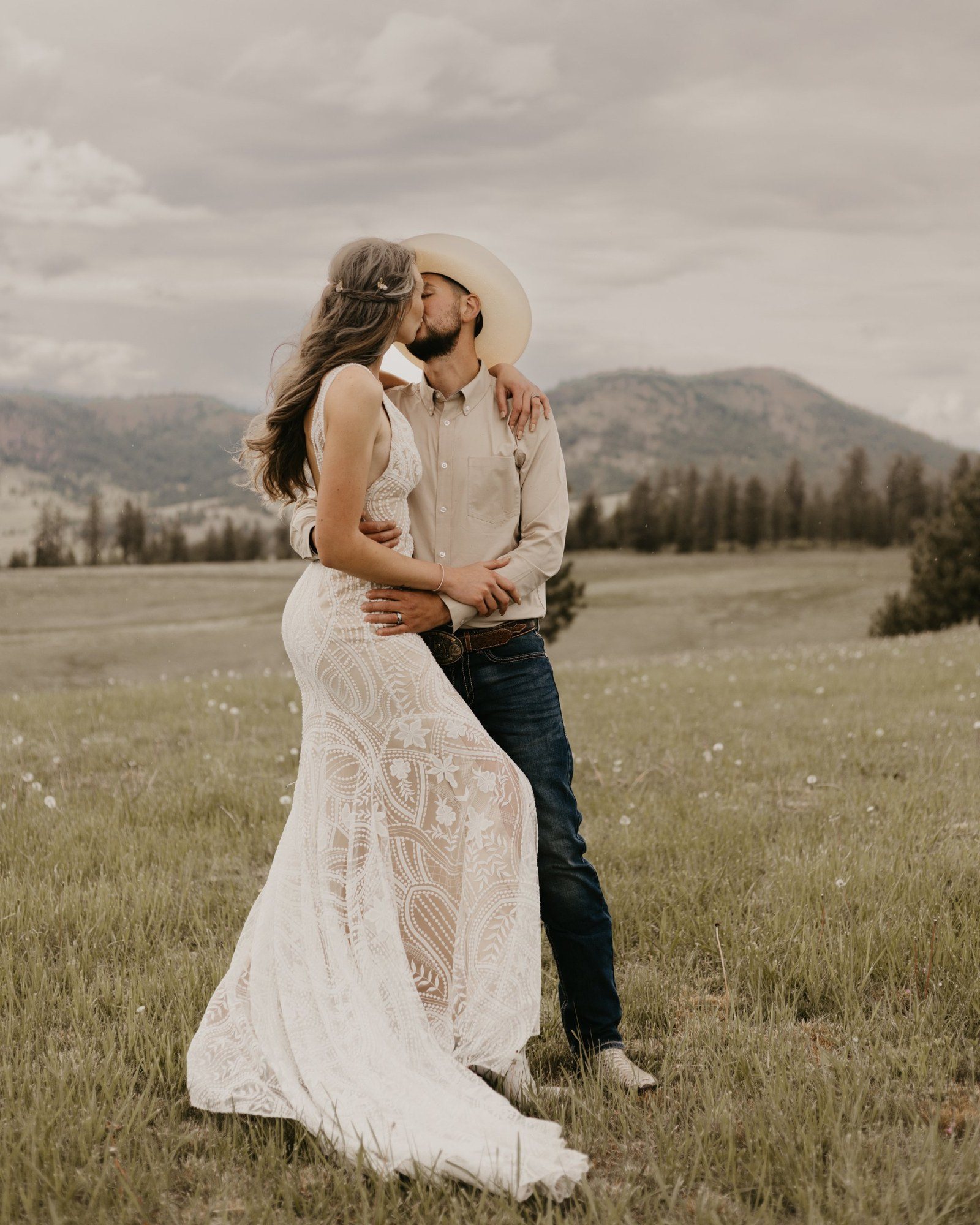Groom in a cowboy hat kissing his bride in a lace gown in a mountain meadow