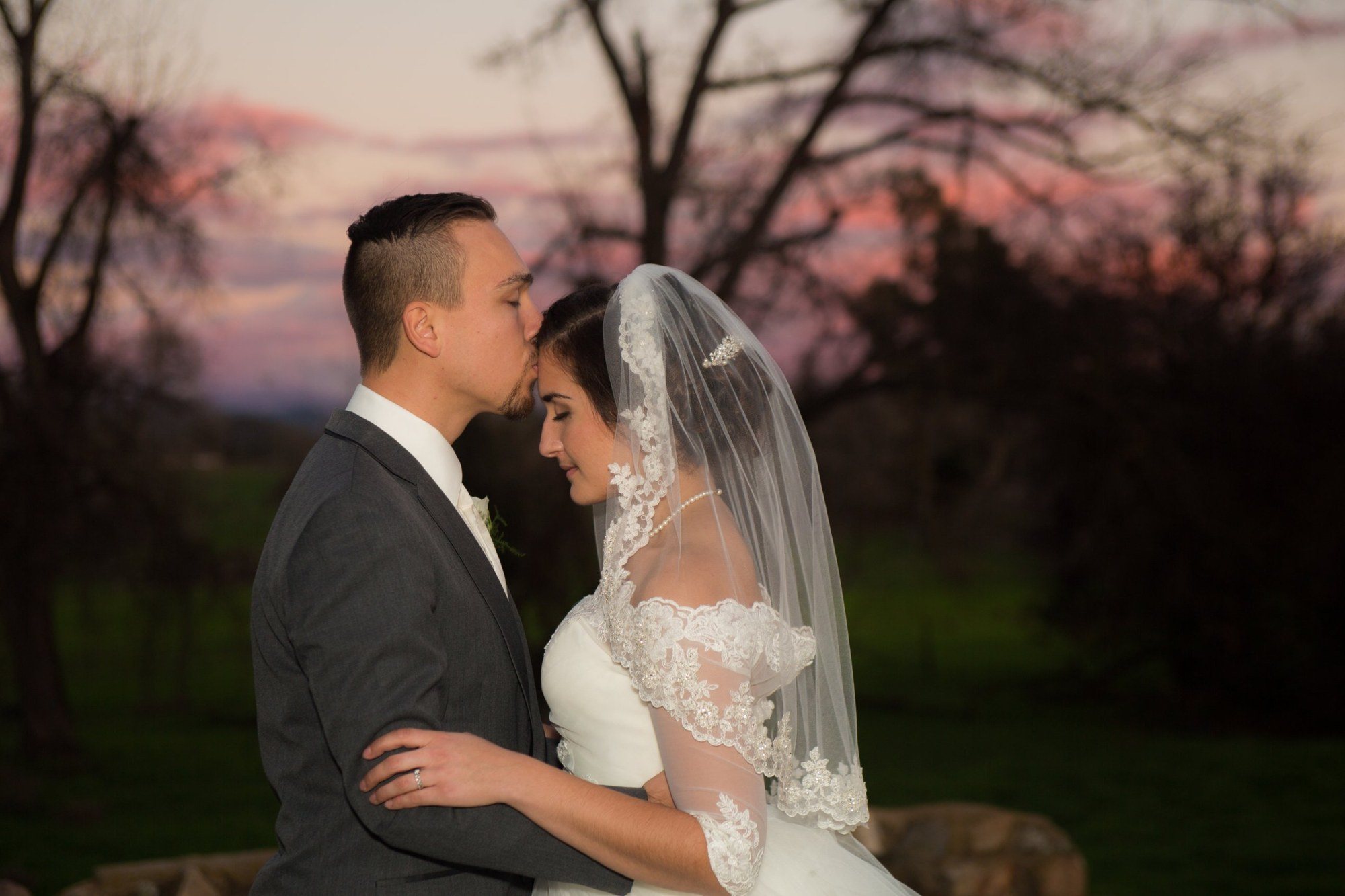 Groom kissing his bride's forehead at dusk under bare trees
