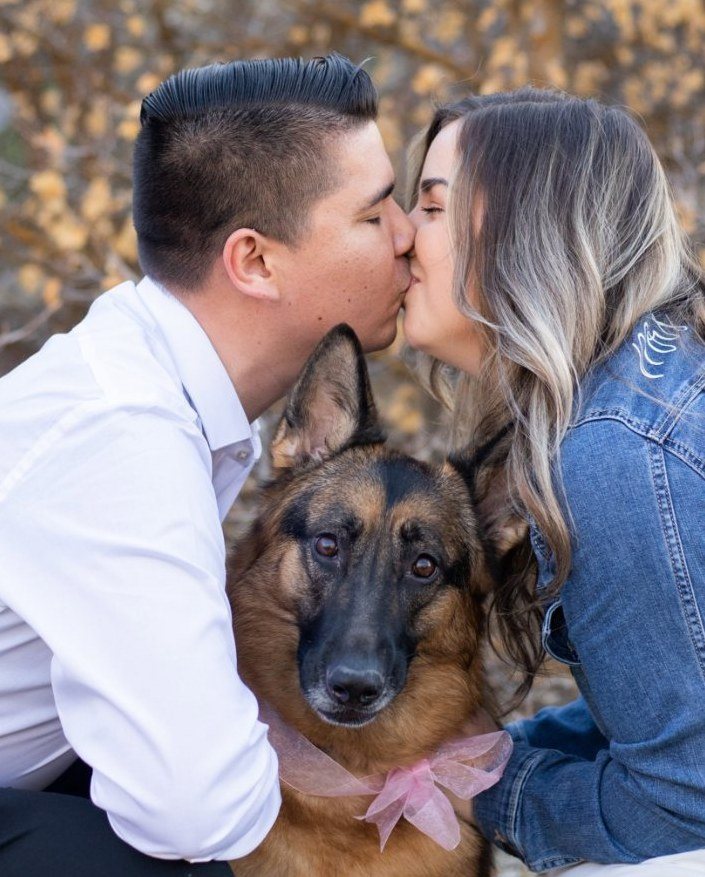 Engaged couple kissing beside their German shepherd