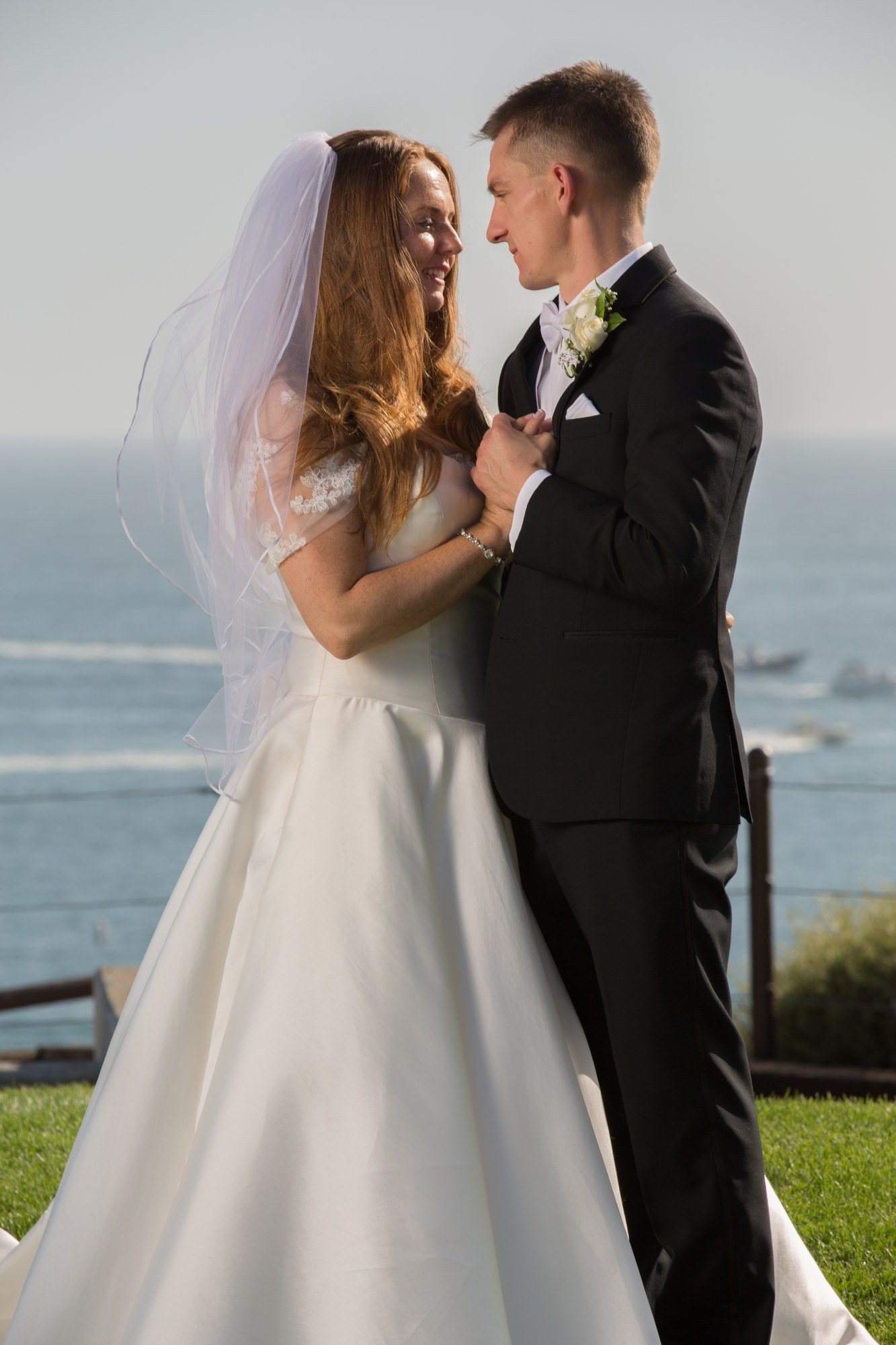 Bride and groom on a lawn above the ocean, veil lifting in the breeze