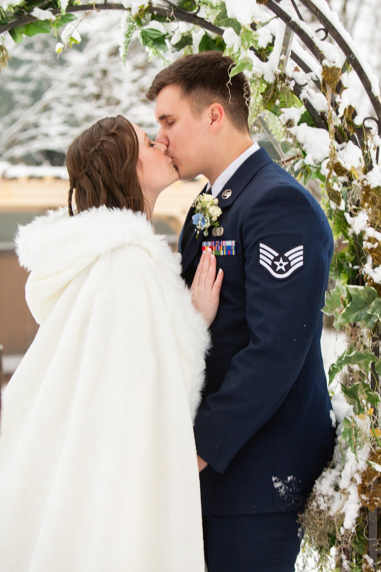 Couple kissing under a snowy floral arch, bride in a white cloak
