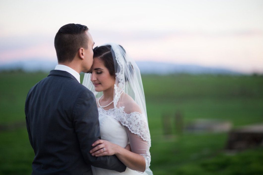 Groom kissing his bride's forehead through her lace veil at dusk